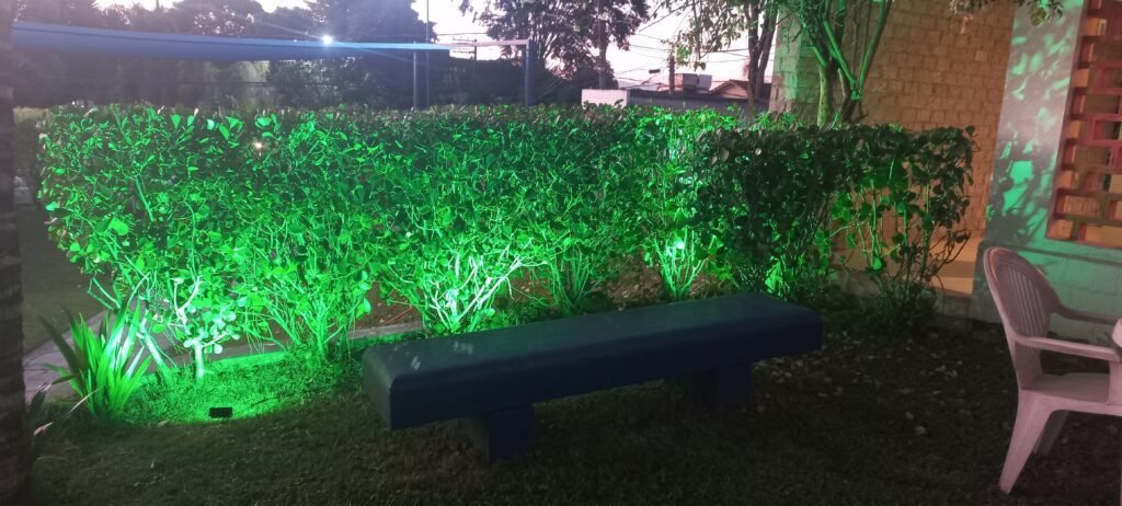 A imagem demonstra um refletor verde iluminando um arbusto em frente a um banco.